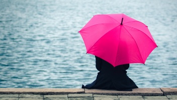Eine in schwarz gekleidete Frau sitzt im Regen an einem Flussufer und hält einen pinken Regenschirm über sich.
