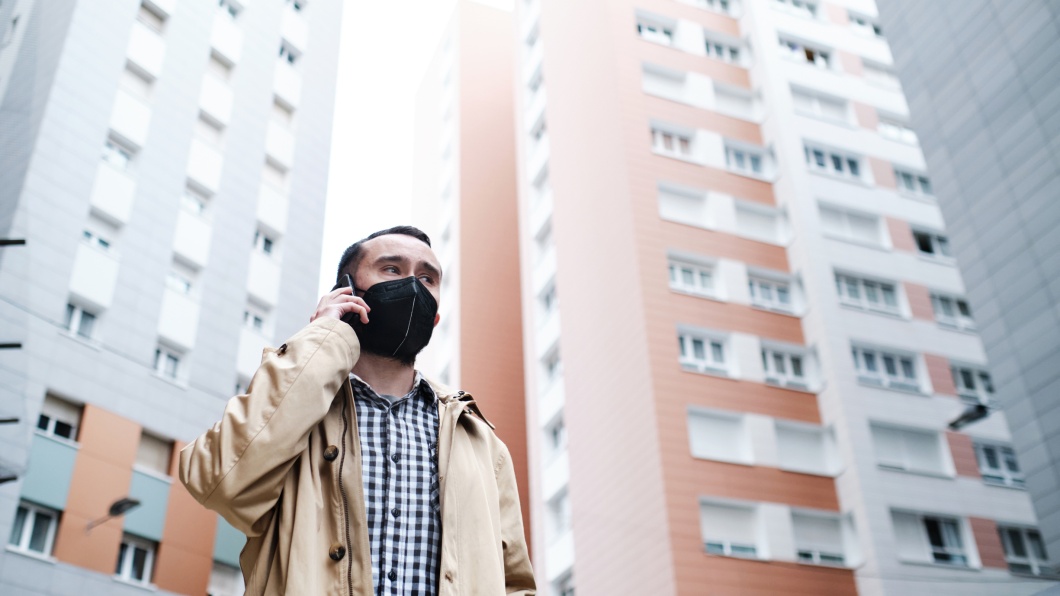 Mann mit Gesichtsschutzmaske, der zwischen hohen Gebäuden mit einem Handy telefoniert