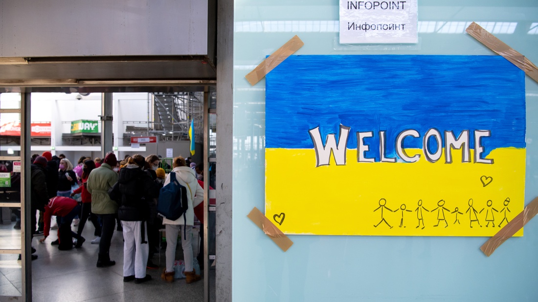 Schild in den Farben der Ukraine mit der Aufschrift "Welcome" am Münchner Hauptbahnhof , im Hintergrund ukrainische Flüchtlinge