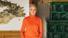 Eine blonde Frau in einem orangen Rollkragenpullover steht in einer Gaststube neben einem grünen Kaminofen