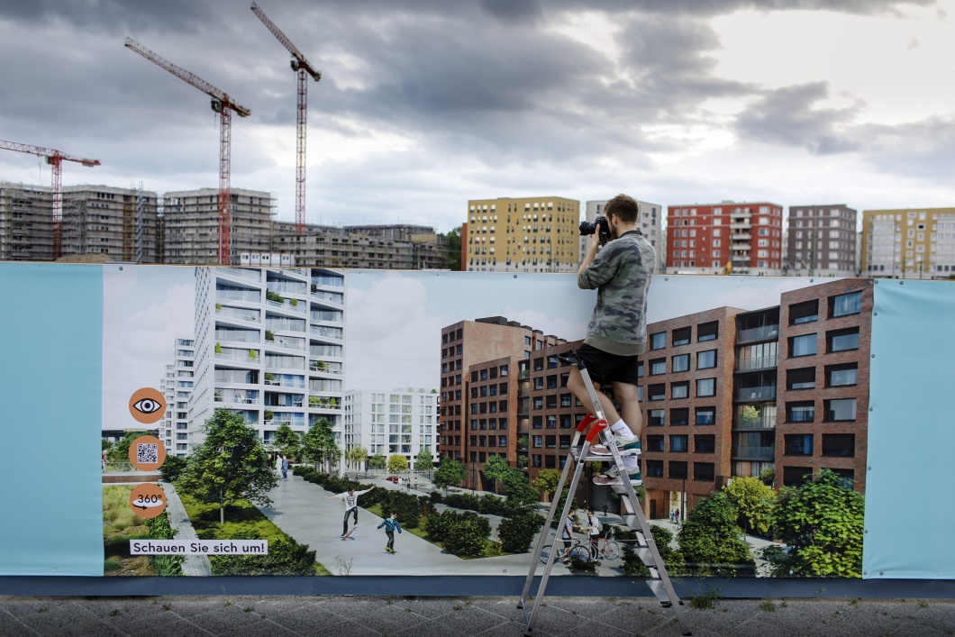 Ein Mann steht auf einer Leiter vor einer Informationswand zum Wohnungsbau und fotografiert eine Baustelle mit Kraenen und Wohnhaeusern im Bau dahinter. 
