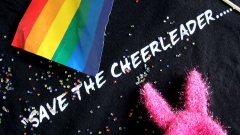 Schwarzes T-Shirt mit dem weißen Schriftzug "Save the Cheerleader", das ist ein Slogan aus der TV-Serie Heroes. Drumherum Glitter und Regenbogenfahne.