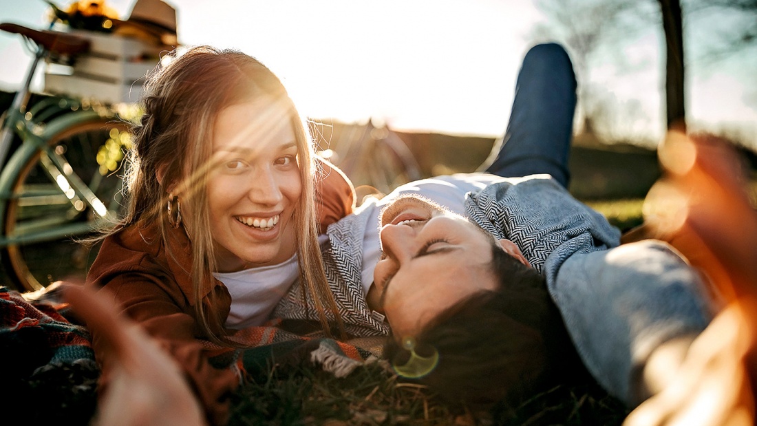 Zwei junge Frauen liegen lachend auf einer Wiese in der Sonne