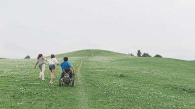 Zwei Frauen helfen einem Mann im Rollstuhl auf einem steilen Weg.