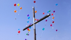 Bunte Luftballons fliegen über einem Holzkreuz in den Himmel.