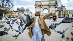 Schwarzer Mensch steht mit erhobenen Armen in Paris vorm Arc de Triomph. Viele Tauben umflattern ihn. 