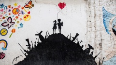 Ein Graffito an einer Wand einer Schule in Hasaka im Nordosten von Syrien zeigt in einem Erdhügel steckende Waffen. Auf der Kuppe stehen zwei Kinder mit einem roten Herz-Luftballon. 